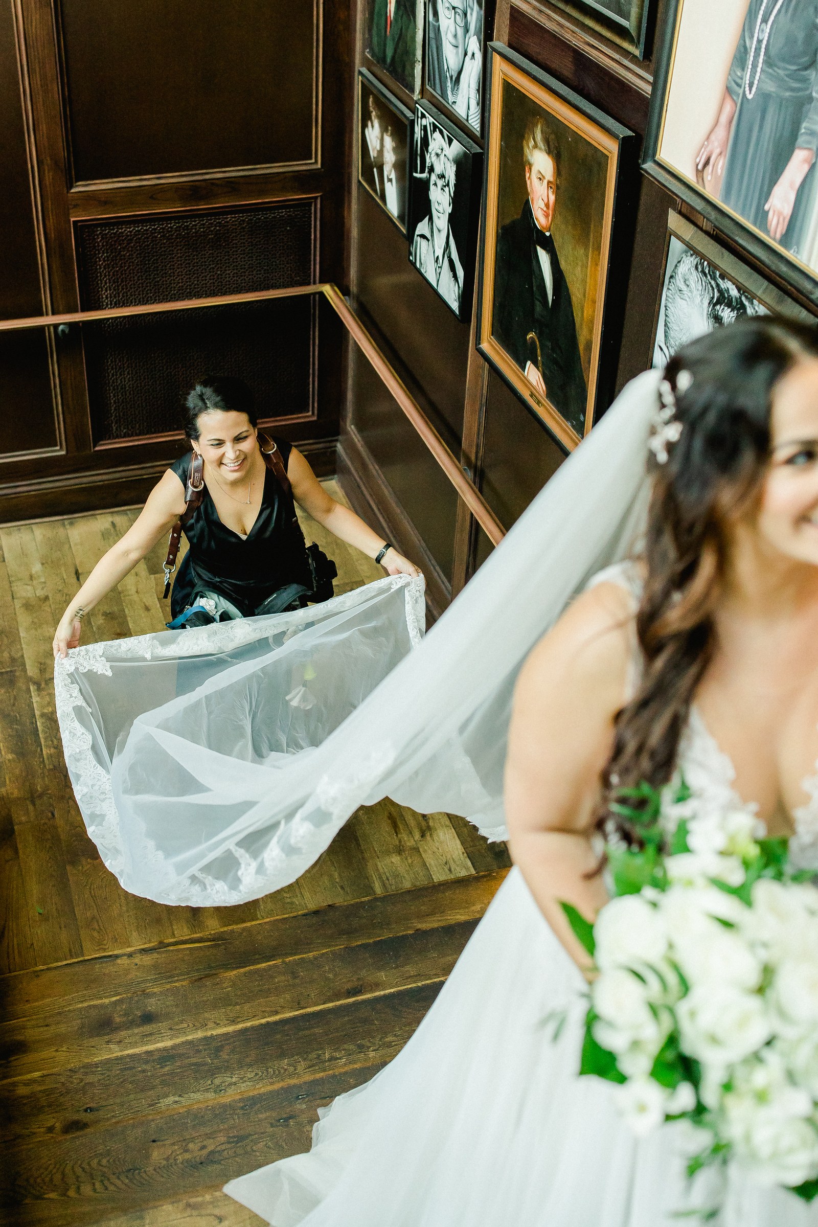 Ailyn La Torre lifting a bride's cathedral veil on the grand staircase at Oxford Exchange in Tampa, dark-wood portrait gallery lining the walls — Tampa Bay wedding photographer
